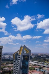 A tall, modern skyscraper with angular architecture stands prominently against a backdrop of a clear blue sky dotted with fluffy white clouds. The building has a shiny glass facade and features the label 'CBK Pension Towers' in gold. Surrounding the structure are smaller buildings and greenery, creating a contrast between urban and natural elements.