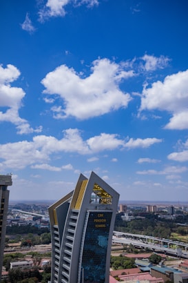 A tall, modern skyscraper with angular architecture stands prominently against a backdrop of a clear blue sky dotted with fluffy white clouds. The building has a shiny glass facade and features the label 'CBK Pension Towers' in gold. Surrounding the structure are smaller buildings and greenery, creating a contrast between urban and natural elements.