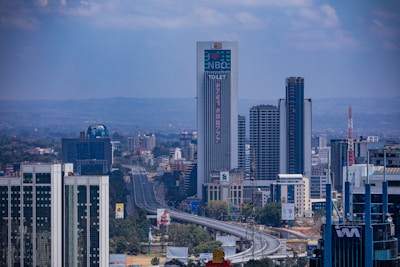 A cityscape featuring tall modern buildings and skyscrapers, with a prominent building in the center displaying the letters 'NBO'. There is a highway running through the scene, leading towards a distant, hazy mountainous horizon. The sky is partly cloudy.
