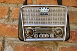 A vintage portable radio with a crown emblem and three stars on its grille, mounted on a red brick wall. The radio features two large knobs for volume and tuning, and a series of buttons and functions labeled on its front panel.