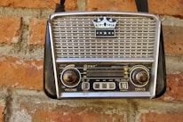 A vintage portable radio with a crown emblem and three stars on its grille, mounted on a red brick wall. The radio features two large knobs for volume and tuning, and a series of buttons and functions labeled on its front panel.