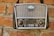 A vintage portable radio with a crown emblem and three stars on its grille, mounted on a red brick wall. The radio features two large knobs for volume and tuning, and a series of buttons and functions labeled on its front panel.