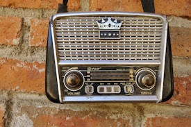 A vintage portable radio with a crown emblem and three stars on its grille, mounted on a red brick wall. The radio features two large knobs for volume and tuning, and a series of buttons and functions labeled on its front panel.