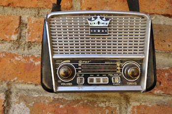 A vintage portable radio with a crown emblem and three stars on its grille, mounted on a red brick wall. The radio features two large knobs for volume and tuning, and a series of buttons and functions labeled on its front panel.