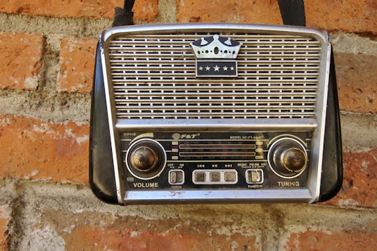 A vintage portable radio with a crown emblem and three stars on its grille, mounted on a red brick wall. The radio features two large knobs for volume and tuning, and a series of buttons and functions labeled on its front panel.