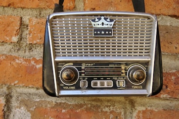 A vintage portable radio with a crown emblem and three stars on its grille, mounted on a red brick wall. The radio features two large knobs for volume and tuning, and a series of buttons and functions labeled on its front panel.