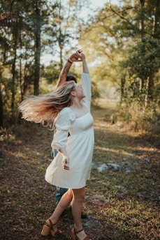 A couple is dancing in a forest setting. The woman is wearing a white dress and brown sandals, with her long hair flowing as she twirls. The man is mostly obscured but appears to be holding her hand, guiding the dance. Sunlight filters through the trees, casting a warm glow on the scene.