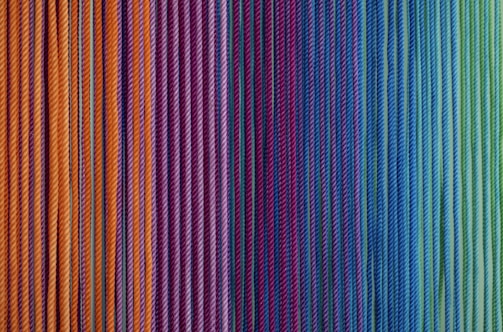 A pattern of tightly packed, colorful yarn strings arranged vertically in a gradient from warm yellows and oranges on the left to cool blues and greens on the right. The texture of the yarns is visible, highlighting their fibrous nature.