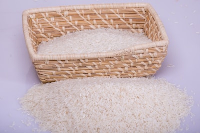 A woven basket is filled with white rice, with some rice spilling over onto a flat surface. The basket is made of a light brown natural material and has a rectangular shape.
