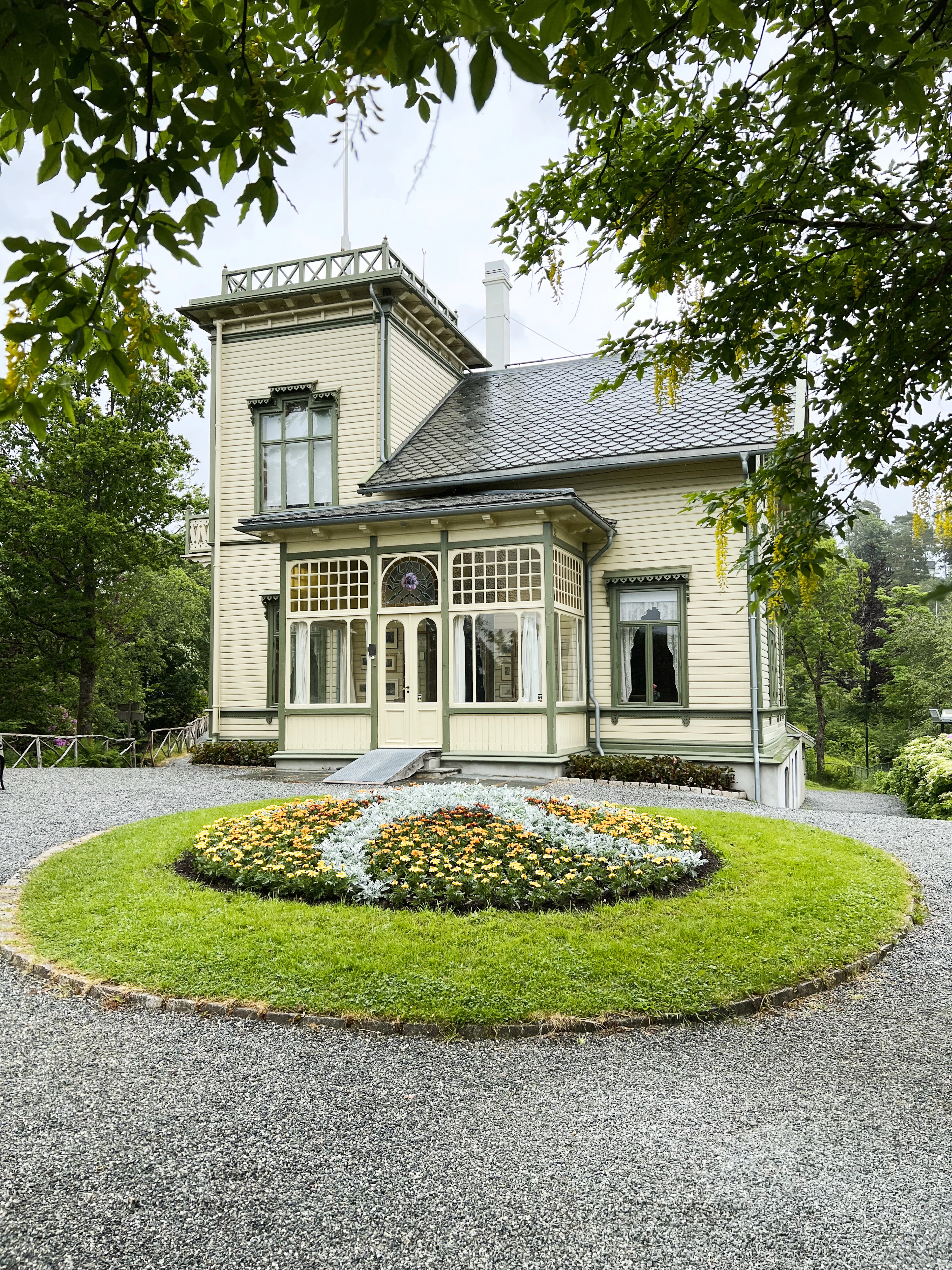 Troldhaugen, Edvard Grieg's pale-yellow timber villa outside Bergen, its glazed verandah and carved gables framed by summer trees above a circular lawn planted with an oval bed of orange and white flowers.