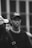 A black and white photograph of a person wearing a baseball cap with the text 'AMIRI' and a graphic on it. The person looks directly at the camera and is dressed in a shirt with 'Acne Studios' on the collar. Another person is partially visible next to them, with focus on the central figure.
