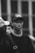 A black and white photograph of a person wearing a baseball cap with the text 'AMIRI' and a graphic on it. The person looks directly at the camera and is dressed in a shirt with 'Acne Studios' on the collar. Another person is partially visible next to them, with focus on the central figure.