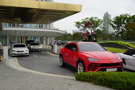 Multiple luxury cars are parked and driving near a modern building with a golden, reflective canopy. The scene includes a vibrant red car in the foreground, a metallic sculpture, and a backdrop of lush green foliage and city skyscrapers. People are walking nearby, adding life to the urban setting.