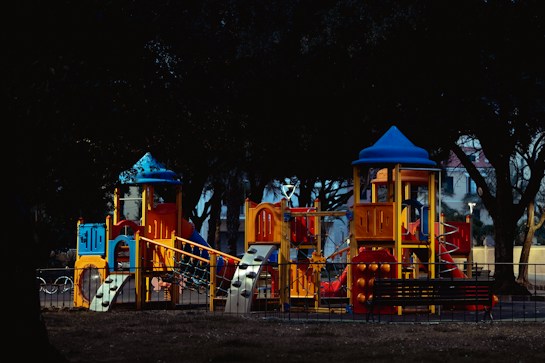 A brightly colored children's playground set is visible, featuring multiple slides, climbing structures, and turrets. The structures are primarily in blue, orange, and yellow hues, standing out against dark trees and a fence surrounding the area. The environment appears calm and quiet, likely at dusk or dawn due to the lighting.