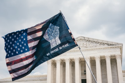A flag design blending the American flag with text forming a raised fist is prominently displayed in front of a neoclassical building with large columns and ornate architectural details.
