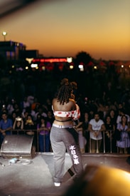 A performer is standing on stage facing a crowd during sunset. The performer is wearing a floral off-shoulder top and dark pants with white symbols on the leg. The audience is gathered closely behind a barrier, with some people taking photos. The sky is a gradient of warm colors from the sunset, and there are building lights in the distance.