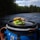 A sandwich with leafy greens and whole grain bread is placed on the lid of a blue container, with a river and dense forest in the background under a partly cloudy sky.