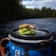A sandwich with leafy greens and whole grain bread is placed on the lid of a blue container, with a river and dense forest in the background under a partly cloudy sky.