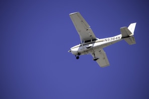 A small aircraft with a registration number visible on the side is flying against a clear blue sky. The airplane's design suggests it is a single-engine plane, likely used for private or recreational flying.