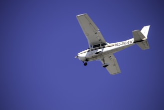A small aircraft with a registration number visible on the side is flying against a clear blue sky. The airplane's design suggests it is a single-engine plane, likely used for private or recreational flying.