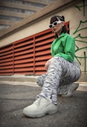 A person is crouching on an urban street, dressed in a vibrant green shirt, zebra-striped pants, and white sneakers. They wear stylish white sunglasses and have their hair styled in cornrows and loose bangs. The background features red industrial vents and a concrete building, with green graffiti visible on the wall.