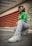 A person is crouching on an urban street, dressed in a vibrant green shirt, zebra-striped pants, and white sneakers. They wear stylish white sunglasses and have their hair styled in cornrows and loose bangs. The background features red industrial vents and a concrete building, with green graffiti visible on the wall.