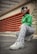 A person is crouching on an urban street, dressed in a vibrant green shirt, zebra-striped pants, and white sneakers. They wear stylish white sunglasses and have their hair styled in cornrows and loose bangs. The background features red industrial vents and a concrete building, with green graffiti visible on the wall.
