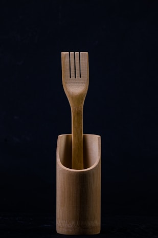 A wooden fork is elegantly placed inside a matching wooden container. The background is dark, emphasizing the natural texture and simplicity of the design.
