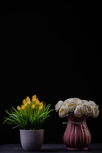 Two flower arrangements are placed side by side against a dark background. The left arrangement features yellow flowers with green foliage in a white pot. The right arrangement consists of white and pink roses in a textured pink vase, tied with a string.