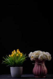 Two flower arrangements are placed side by side against a dark background. The left arrangement features yellow flowers with green foliage in a white pot. The right arrangement consists of white and pink roses in a textured pink vase, tied with a string.
