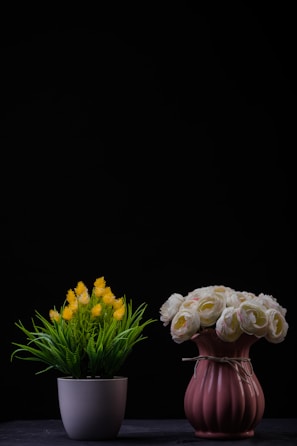 Two flower arrangements are placed side by side against a dark background. The left arrangement features yellow flowers with green foliage in a white pot. The right arrangement consists of white and pink roses in a textured pink vase, tied with a string.