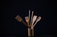 A collection of wooden kitchen utensils is neatly arranged in a cylindrical holder against a dark background. The utensils include a spatula, a slotted spatula, a spoon, and a pair of chopsticks. The natural wood grain and texture are visible, and the composition is minimalistic, emphasizing the simplicity of the wooden tools.