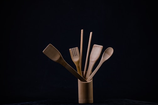 A collection of wooden kitchen utensils is neatly arranged in a cylindrical holder against a dark background. The utensils include a spatula, a slotted spatula, a spoon, and a pair of chopsticks. The natural wood grain and texture are visible, and the composition is minimalistic, emphasizing the simplicity of the wooden tools.