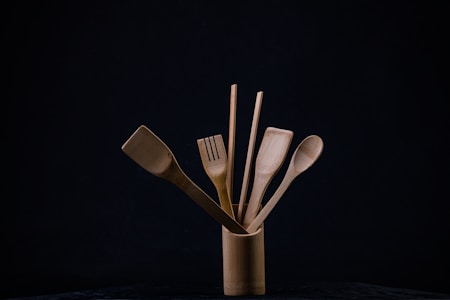 A collection of wooden kitchen utensils is neatly arranged in a cylindrical holder against a dark background. The utensils include a spatula, a slotted spatula, a spoon, and a pair of chopsticks. The natural wood grain and texture are visible, and the composition is minimalistic, emphasizing the simplicity of the wooden tools.