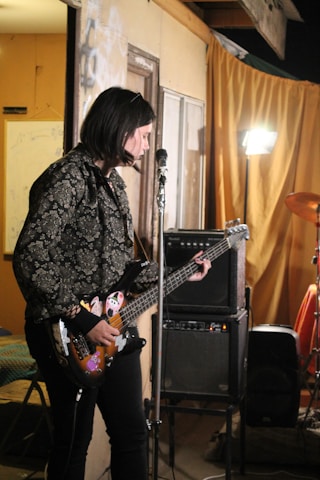 A person is playing an electric bass guitar with stickers on it in a room. There is an amplifier in the background along with a drum set partially visible. A yellow-orange curtain and a bright light stand out in the background, creating an intimate, possibly rehearsal or casual performance setting.