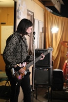 A person is playing an electric bass guitar with stickers on it in a room. There is an amplifier in the background along with a drum set partially visible. A yellow-orange curtain and a bright light stand out in the background, creating an intimate, possibly rehearsal or casual performance setting.