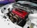 A close-up view of a car's engine bay, featuring a bright red valve cover and complex exhaust manifold. The engine is clean and appears to be modified for high performance, with various metal components and tubing visible. The surrounding car body is white, and the words 'IMPORT LIFE' are displayed on the windshield.
