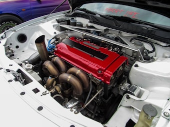 A close-up view of a car's engine bay, featuring a bright red valve cover and complex exhaust manifold. The engine is clean and appears to be modified for high performance, with various metal components and tubing visible. The surrounding car body is white, and the words 'IMPORT LIFE' are displayed on the windshield.