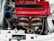 A high-performance car engine is shown, featuring a prominent red VTEC valve cover. The engine components, including a detailed exhaust manifold and various hoses and wires, are visible. The setup appears clean and well-maintained, suggesting it's a part of a modified vehicle built for speed.