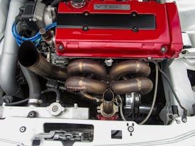 A high-performance car engine is shown, featuring a prominent red VTEC valve cover. The engine components, including a detailed exhaust manifold and various hoses and wires, are visible. The setup appears clean and well-maintained, suggesting it's a part of a modified vehicle built for speed.