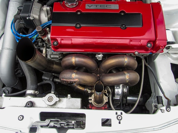 A high-performance car engine is shown, featuring a prominent red VTEC valve cover. The engine components, including a detailed exhaust manifold and various hoses and wires, are visible. The setup appears clean and well-maintained, suggesting it's a part of a modified vehicle built for speed.