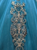 Intricate floral embroidery adorns a teal fabric, featuring gold and shimmering blue accents.