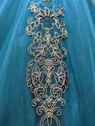 Intricate floral embroidery adorns a teal fabric, featuring gold and shimmering blue accents.