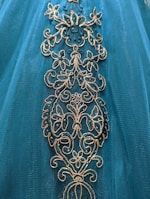 Intricate floral embroidery adorns a teal fabric, featuring gold and shimmering blue accents.