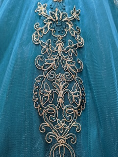 Intricate floral embroidery adorns a teal fabric, featuring gold and shimmering blue accents.