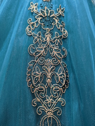 Intricate floral embroidery adorns a teal fabric, featuring gold and shimmering blue accents.
