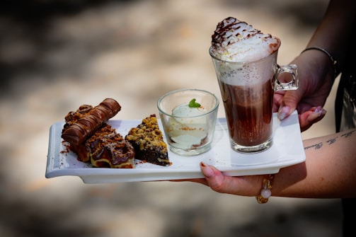 A person is holding a white tray that contains an array of desserts, including a slice of chocolate cake topped with nuts, a cream-filled chocolate roll, and a small glass cup with a mint garnish. Accompanying these is a tall glass of hot chocolate or coffee topped with whipped cream and cocoa powder.