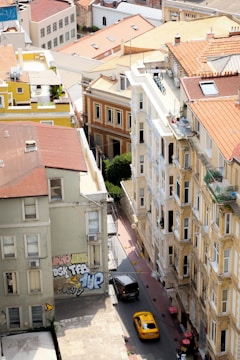 Aerial view of an urban area featuring several multi-story buildings with varied architectural designs. The buildings have different colored facades, including beige, yellow, and red tones. A narrow street runs between them with a yellow taxi and a dark-colored vehicle. Graffiti is visible on the wall of one building.