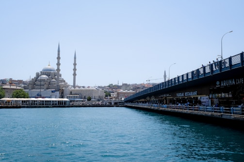 A scenic view of a cityscape featuring a large mosque with multiple domes and two tall minarets on the left side, and a bustling bridge with various cafes and restaurants beneath it on the right side. The foreground shows a calm body of water reflecting the city.