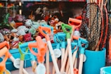 A vibrant collection of beach toys including shovels with colorful handles and nets in a bucket, displayed alongside a variety of plush toys inside a shop window.